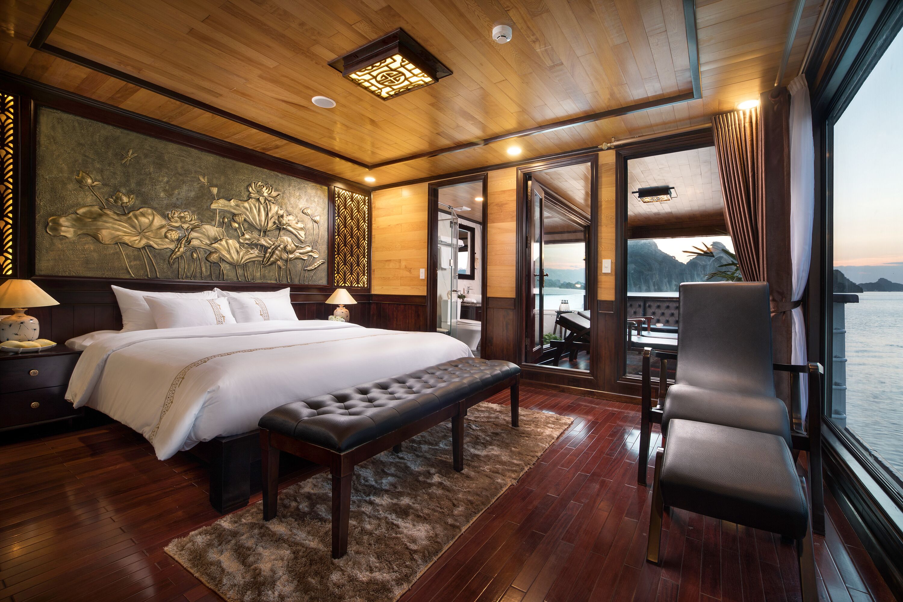 A spacious bedroom with a large bed, a bench, a chair, and a view of the water through large windows.