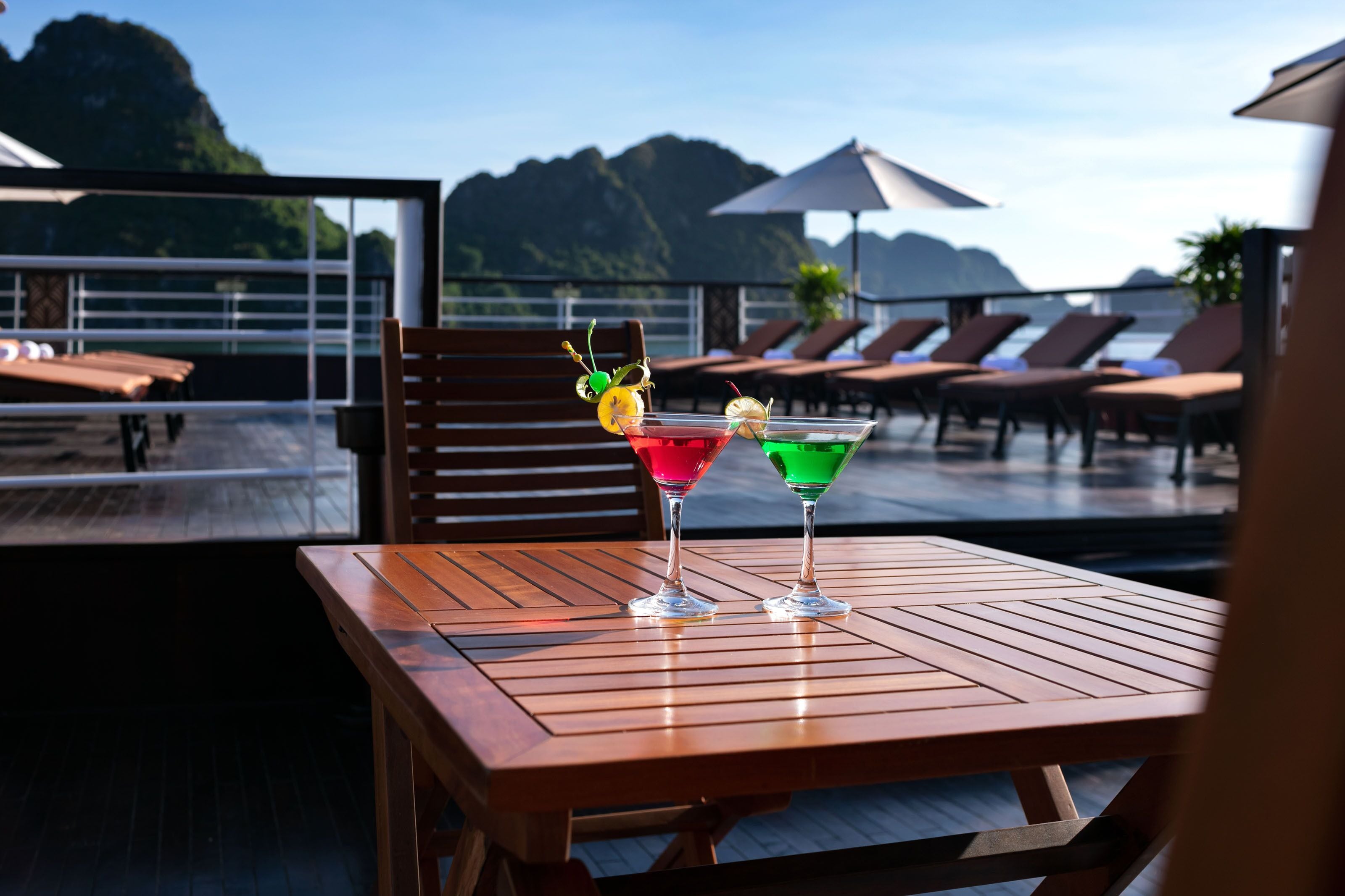 Ein Holztisch mit zwei farbenfrohen Cocktails, Blick auf einen Poolbereich mit Liegestühlen und Sonnenschirmen.