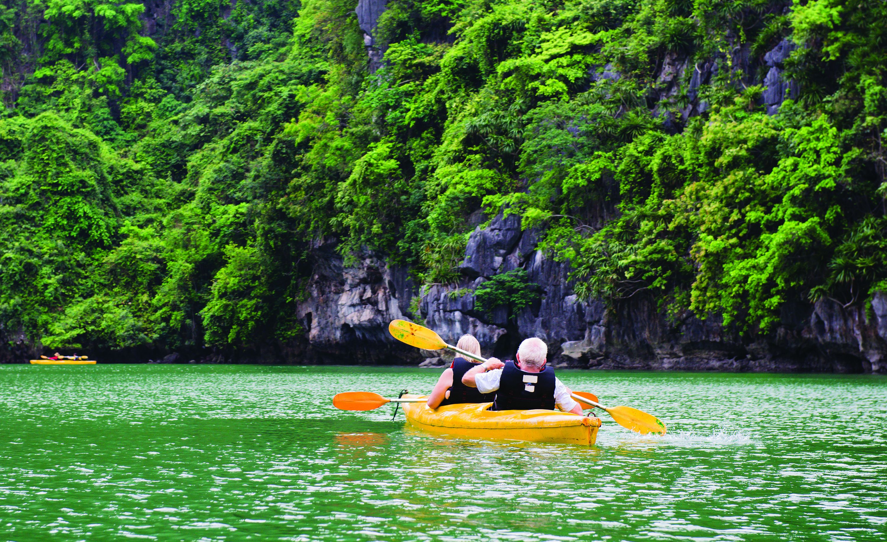 A kayak with two people paddling on a greenish water body surrounded by dense green foliage and rocky cliffs.