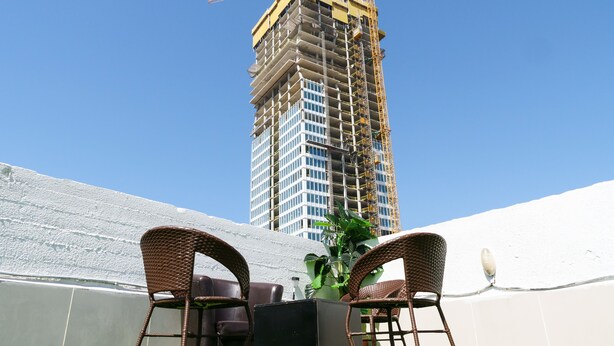 Een hoog gebouw in aanbouw met een kraan, een witte muur, en twee rieten stoelen met een zwarte tafel op de voorgrond.