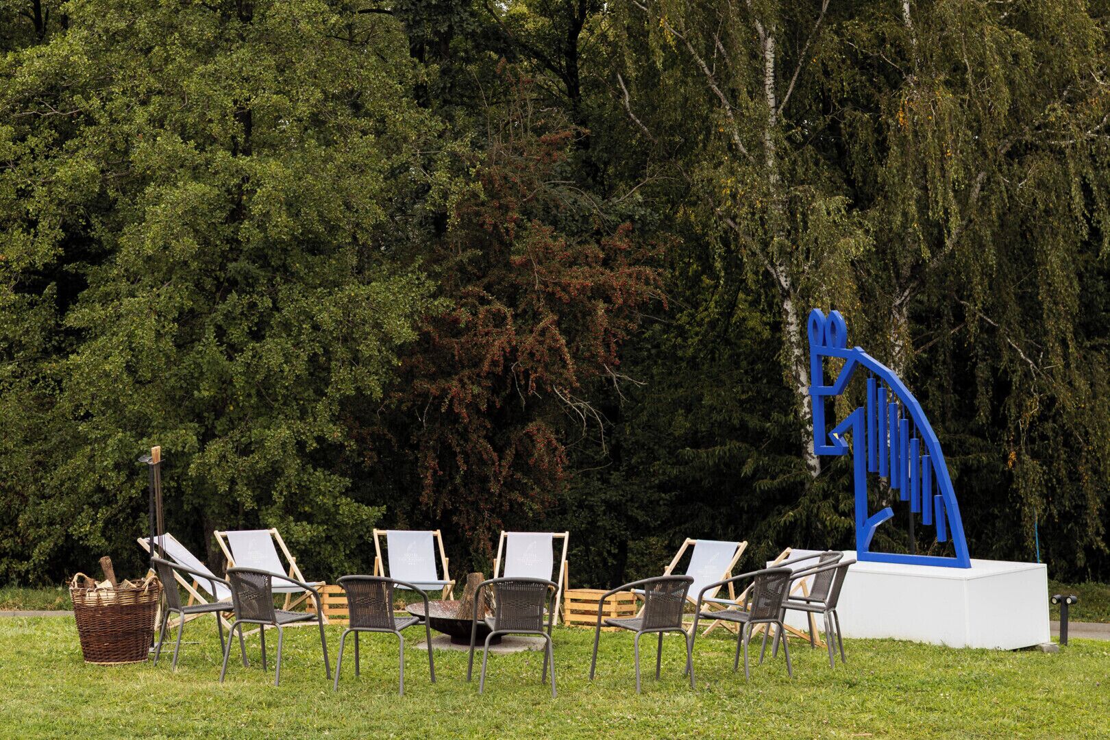 Uma área externa com mesas e cadeiras, uma escultura azul e um fundo arborizado.