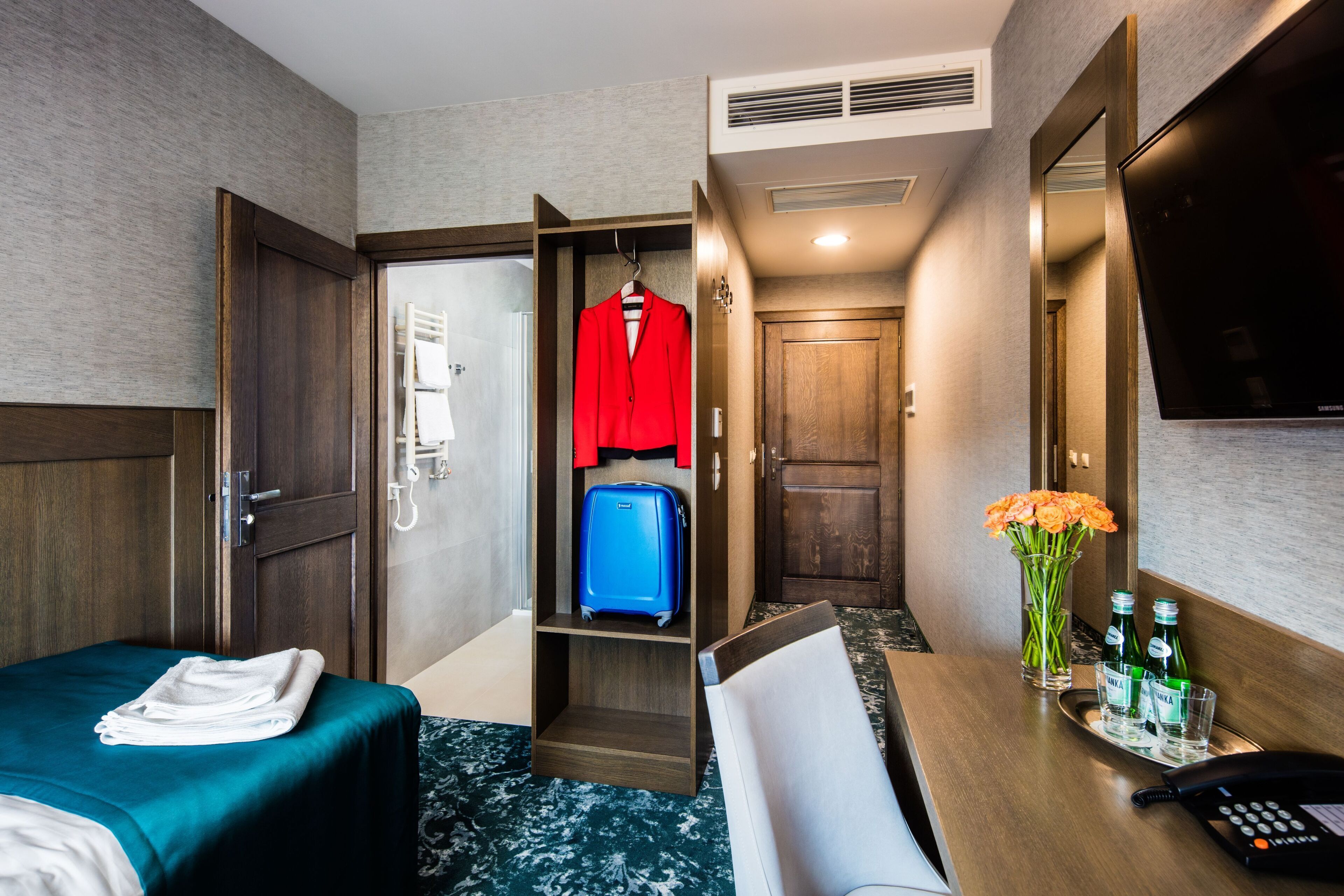 Una habitación de hotel con una cama, un escritorio con teléfono, un armario que contiene una chaqueta roja y una maleta azul, y un jarrón con flores.