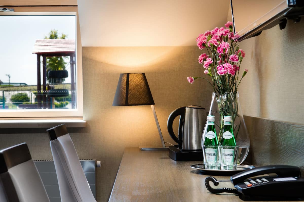 Quarto de hotel com uma mesa de madeira, duas cadeiras, um vaso com flores cor-de-rosa, um abajur e uma chaleira.