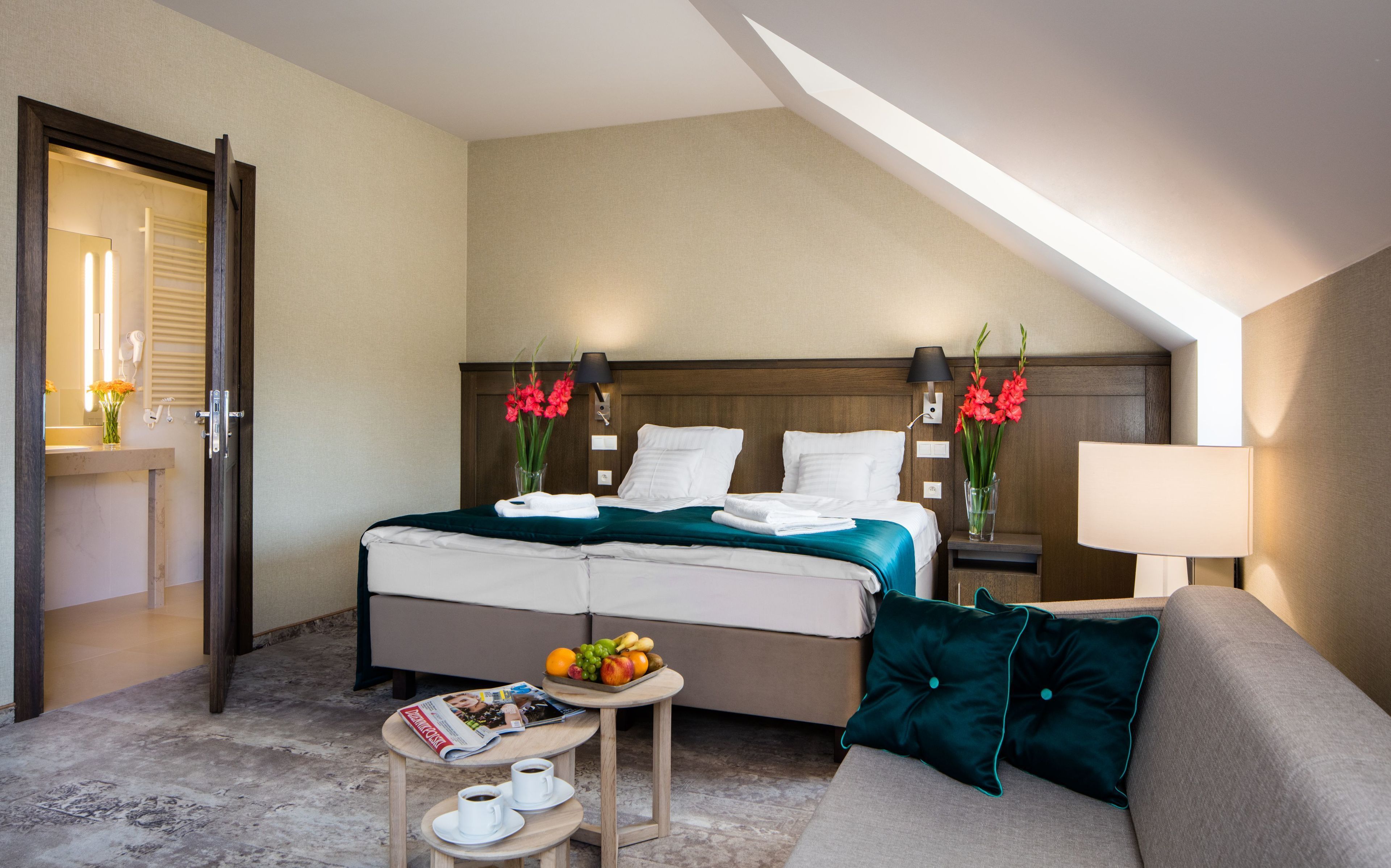 Habitación de hotel con una cama, una zona de estar con sofá, una mesita con frutero y revista, y un baño visible a través de una puerta abierta.