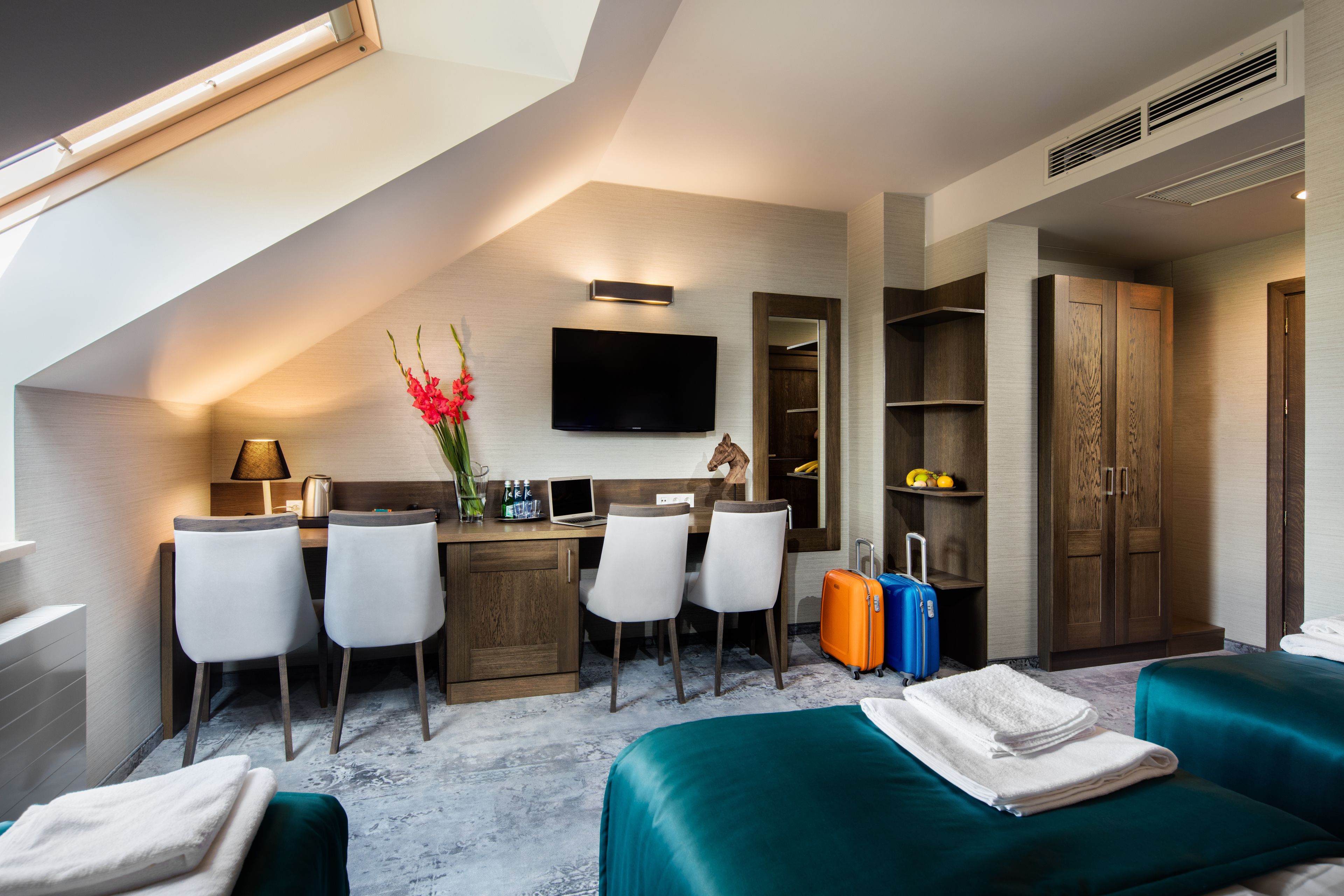 Una habitación de hotel moderna con techo estilo loft, un escritorio con sillas, un televisor de pantalla plana, un armario y dos camas.