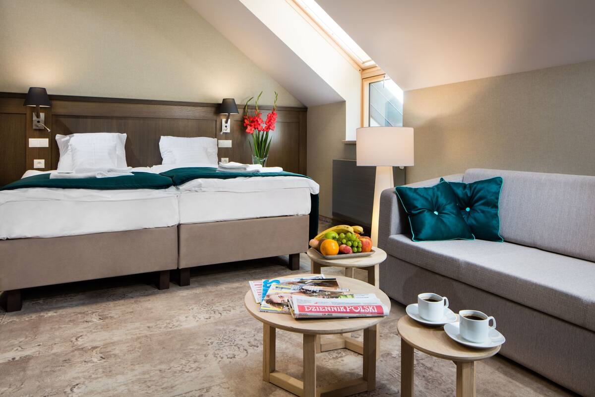 Quarto de hotel com uma cama, um sofá, duas mesinhas, uma luminária e uma janela com luz natural.