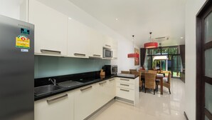 A modern kitchen with white cabinets, a stainless steel refrigerator, and a microwave. There is a dining area with wicker chairs and a red pendant light. - Saiyuan Estate by TropicLook (Rawai)
