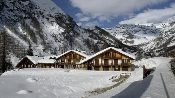 Un hôtel de style chalet, composé de plusieurs cabanes en bois, situé dans un cadre magnifique avec des montagnes enneigées en toile de fond et un ciel dégagé.