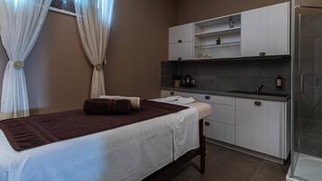 A bedroom with a bed, a small bedside table, a window with curtains, a kitchenette with cabinets, and a shower area.