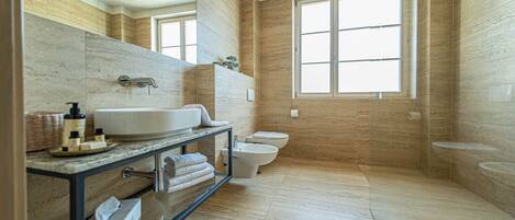 Un baño moderno con un lavamanos grande, inodoro y bidé. Cuenta con una pared de mármol, un ventanal amplio y una cabina de ducha con paredes de cristal.