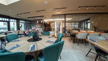 A modern restaurant with round tables and teal chairs, featuring a large vase of flowers and a view of the street through large windows.