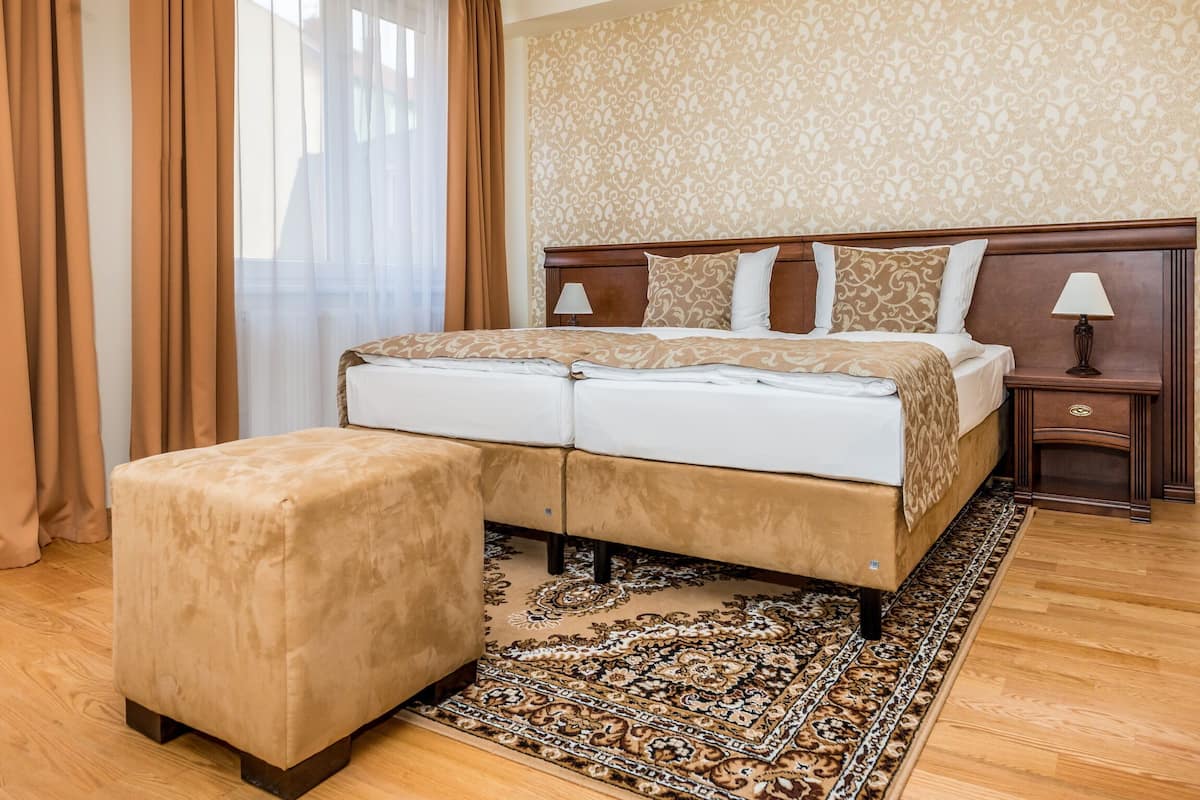 A hotel room with a bed, a nightstand, a lamp, a large ottoman, and a patterned rug.