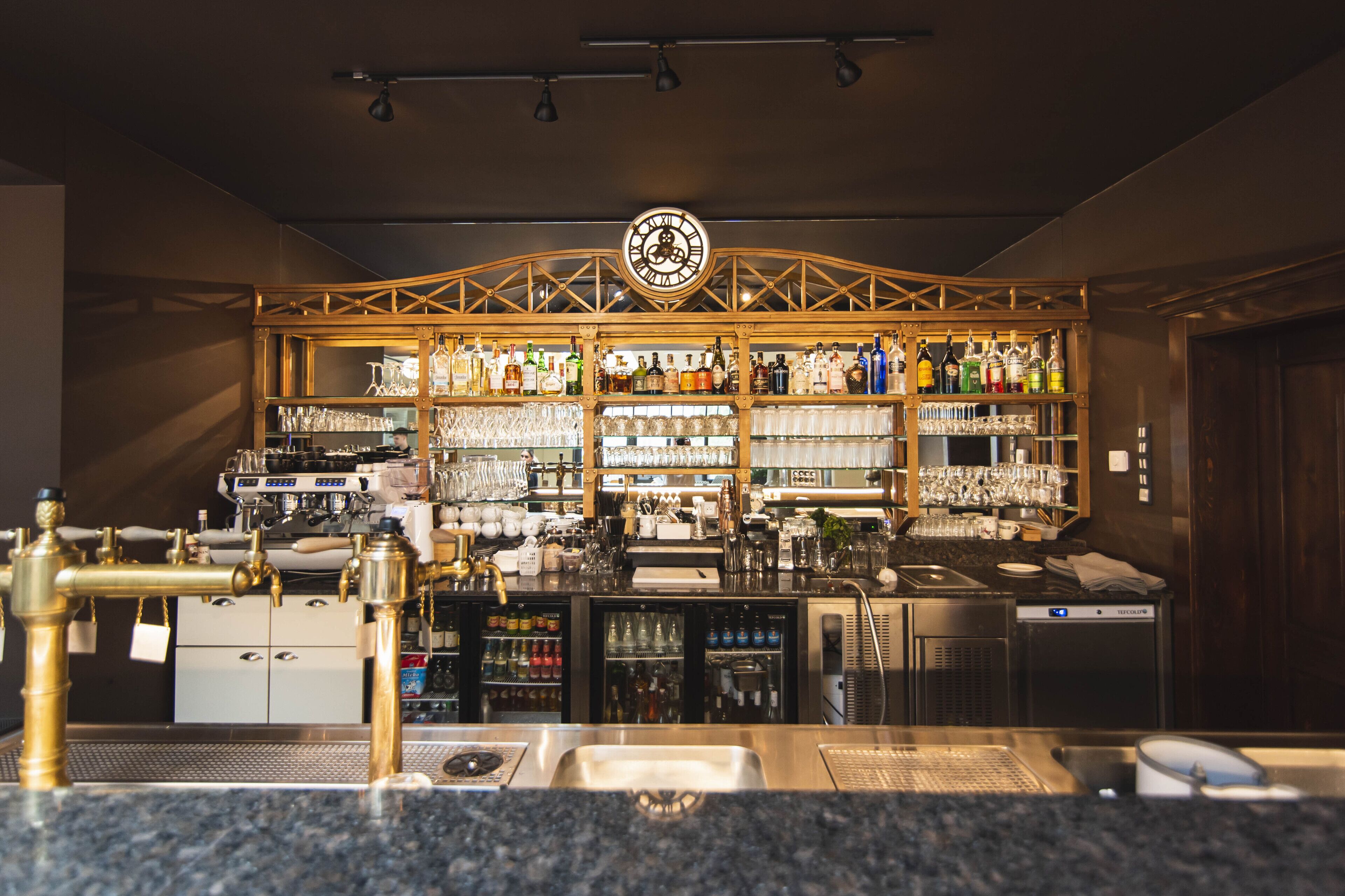 Una zona de bar con un reloj, estantes llenos de botellas, vasos y una cafetera.