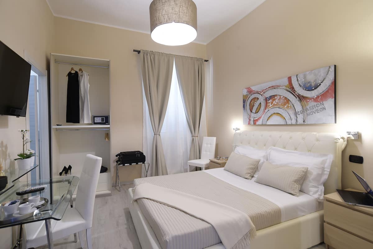 A hotel room with a bed, a desk with a laptop, a chair, a wardrobe, and a painting on the wall.