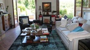 A living room with a sofa, armchair, coffee table, and books. There is a fireplace, a mirror, and a window with a view of greenery. - Donna Etrusca B&B (Sutri)