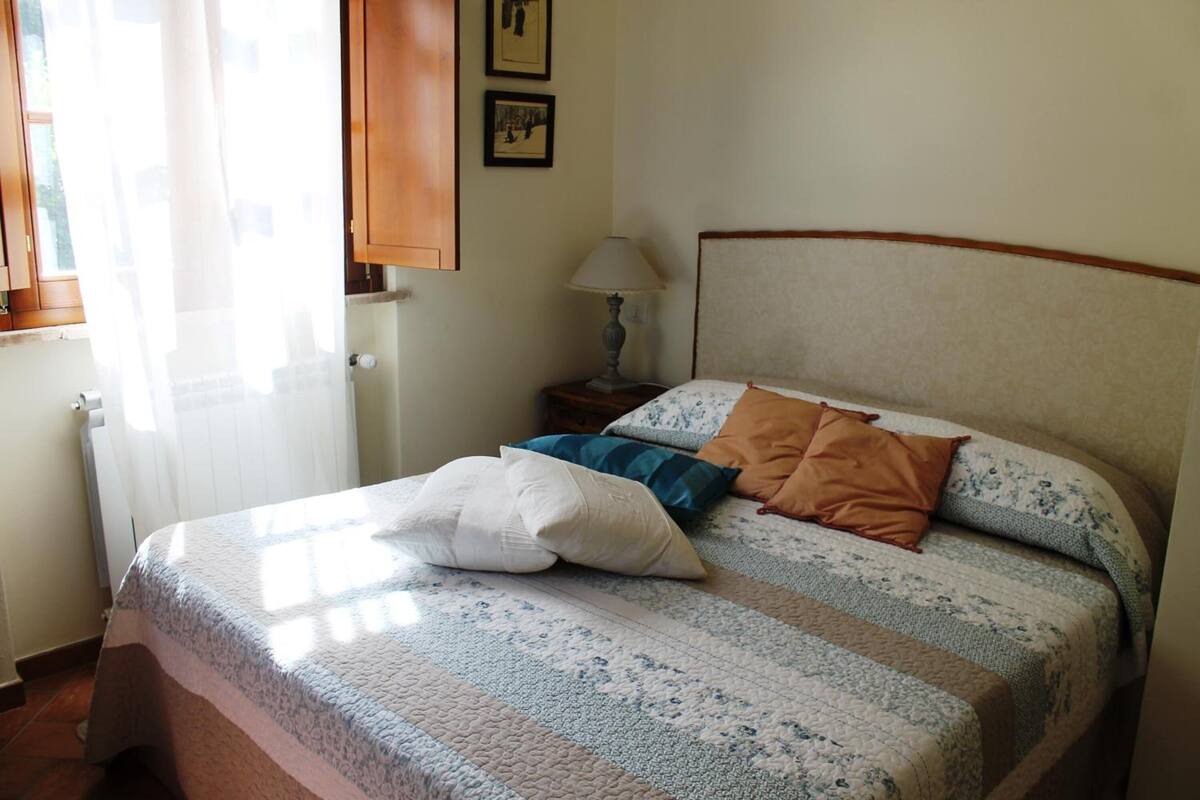 Ein Schlafzimmer mit einem Bett, Kissen, einem Nachttisch, einer Lampe und einem Fenster mit Vorhängen.