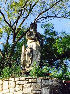 A stone statue of a figure on a pedestal, surrounded by greenery and trees.