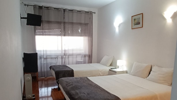 A hotel room with two beds, a desk with a chair, a radiator, and a window with sheer curtains. - Residencial S. Gião (Valenca)