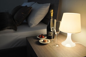 A neatly made bed with pillows, a bottle of champagne, two glasses, and a plate with strawberries and a pastry on a bedside table.