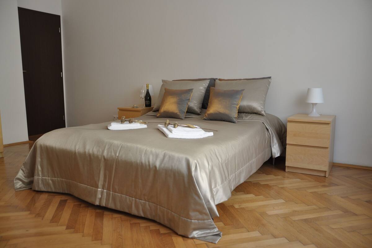 A neatly made bed with a beige bedspread, two pillows, and a nightstand with a lamp and a bottle.