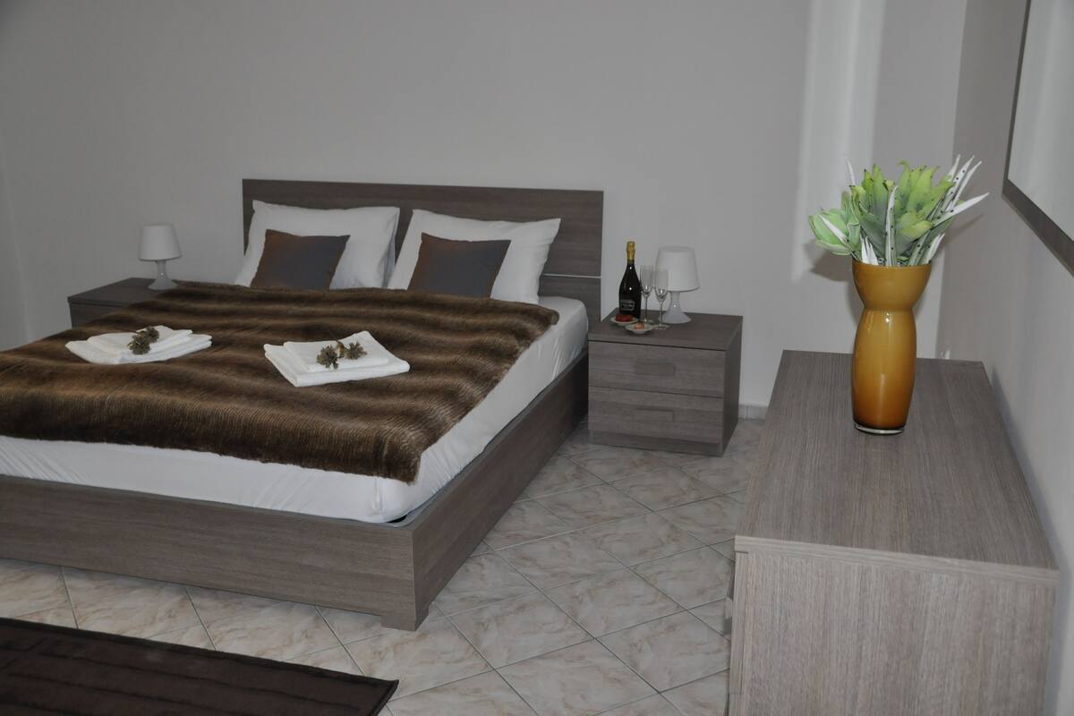 A neatly made bed with a brown blanket, a bedside table with a bottle and glasses, and a vase with green plants.