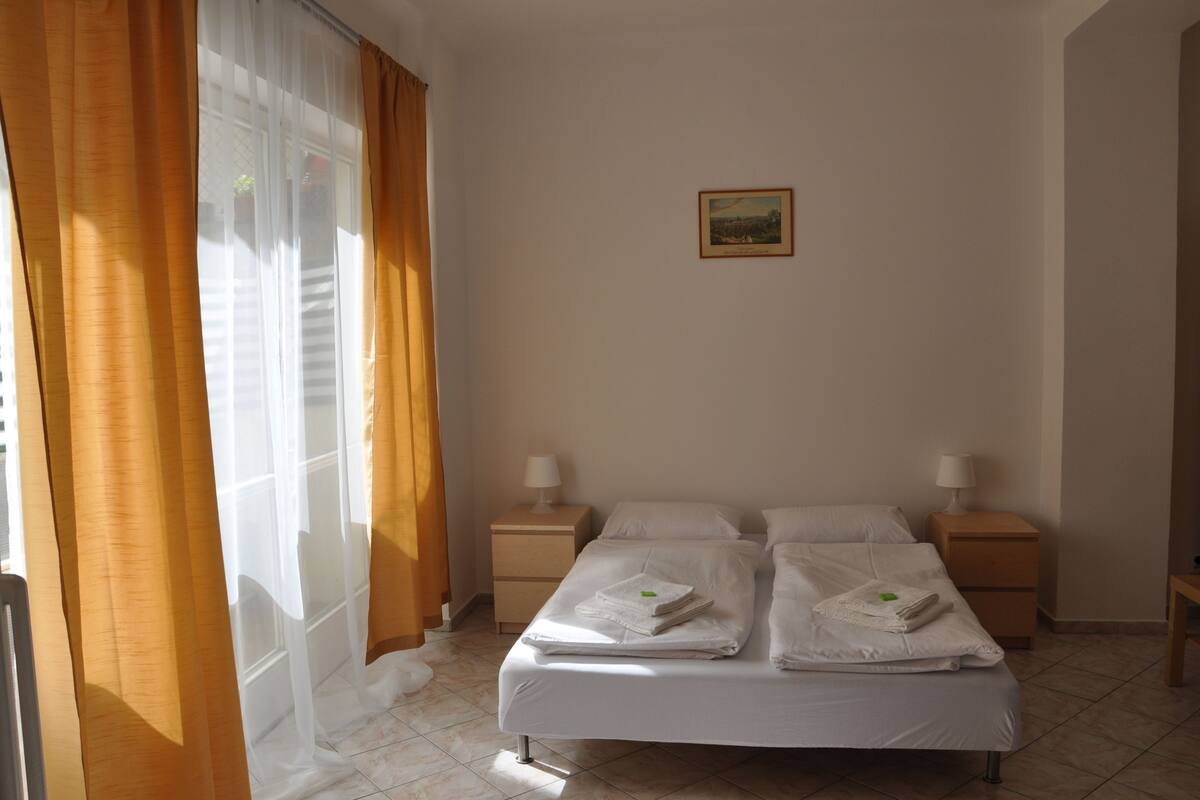 A neatly made bed with white linens and towels, two bedside tables with lamps, and a window with curtains.