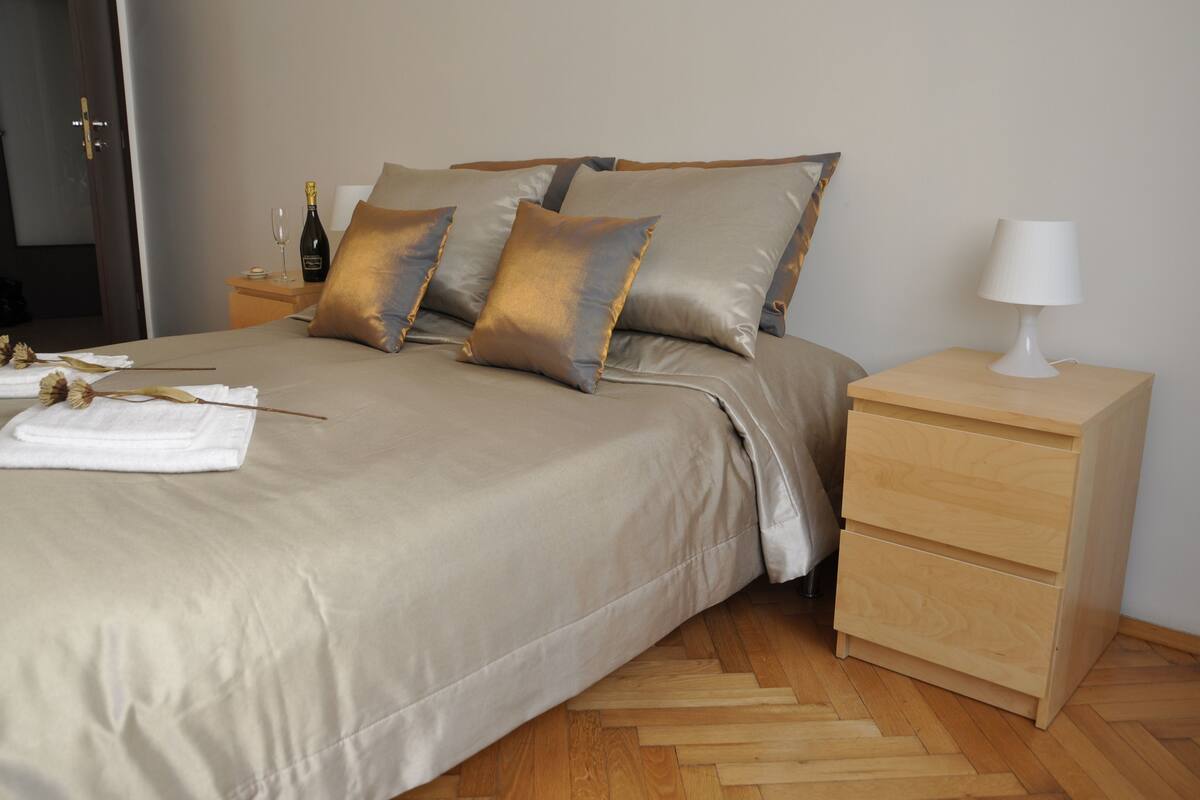 A neatly made bed with a beige comforter and pillows, a bedside table with a lamp, and a wooden floor.