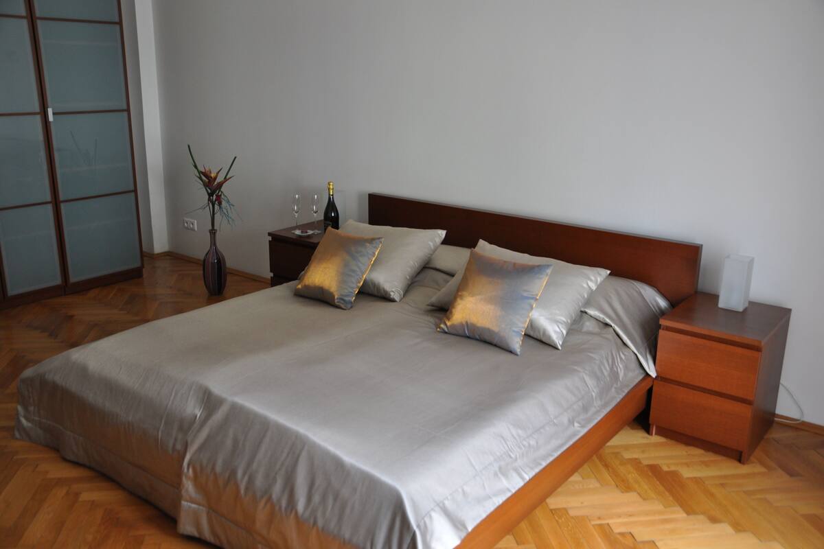 A neatly made bed with a wooden headboard, a bedside table with a vase and a bottle, and a herringbone wood floor.