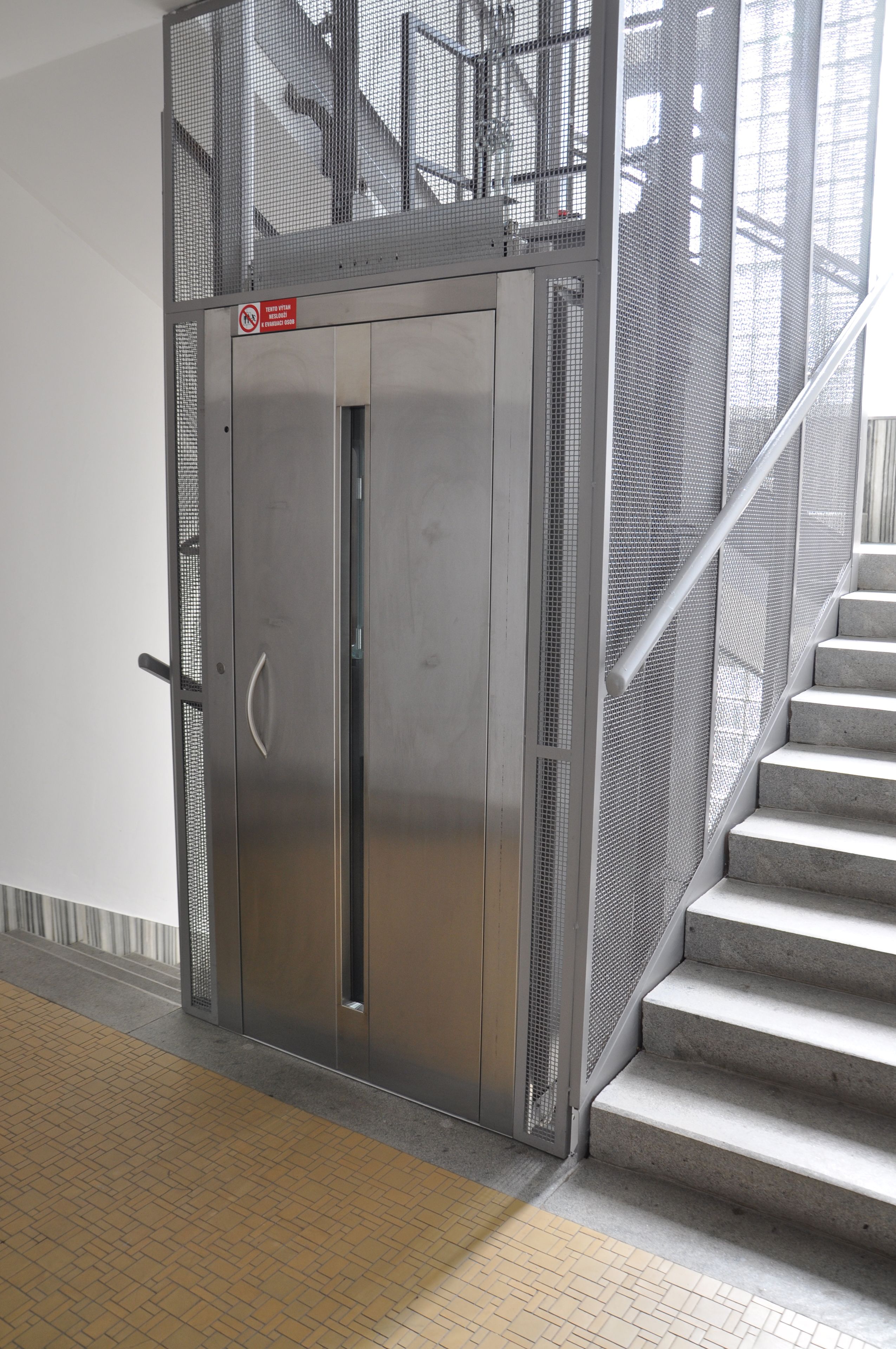 Ein metallener Aufzug mit einem Drahtgitter-Schutzgitter neben einer Treppe mit Metallgeländer.