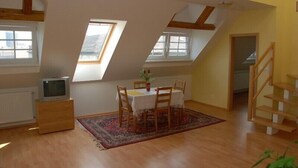A room with a sloped ceiling, wooden beams, a dining table with chairs, a small TV on a dresser, and a staircase. - Apartmany Bratislava (Bratislava)