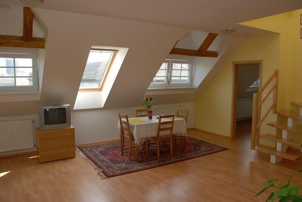 Een kamer met een schuine balken plafond, een eettafel met stoelen, een kleine televisie op een dressoir, en een trap.