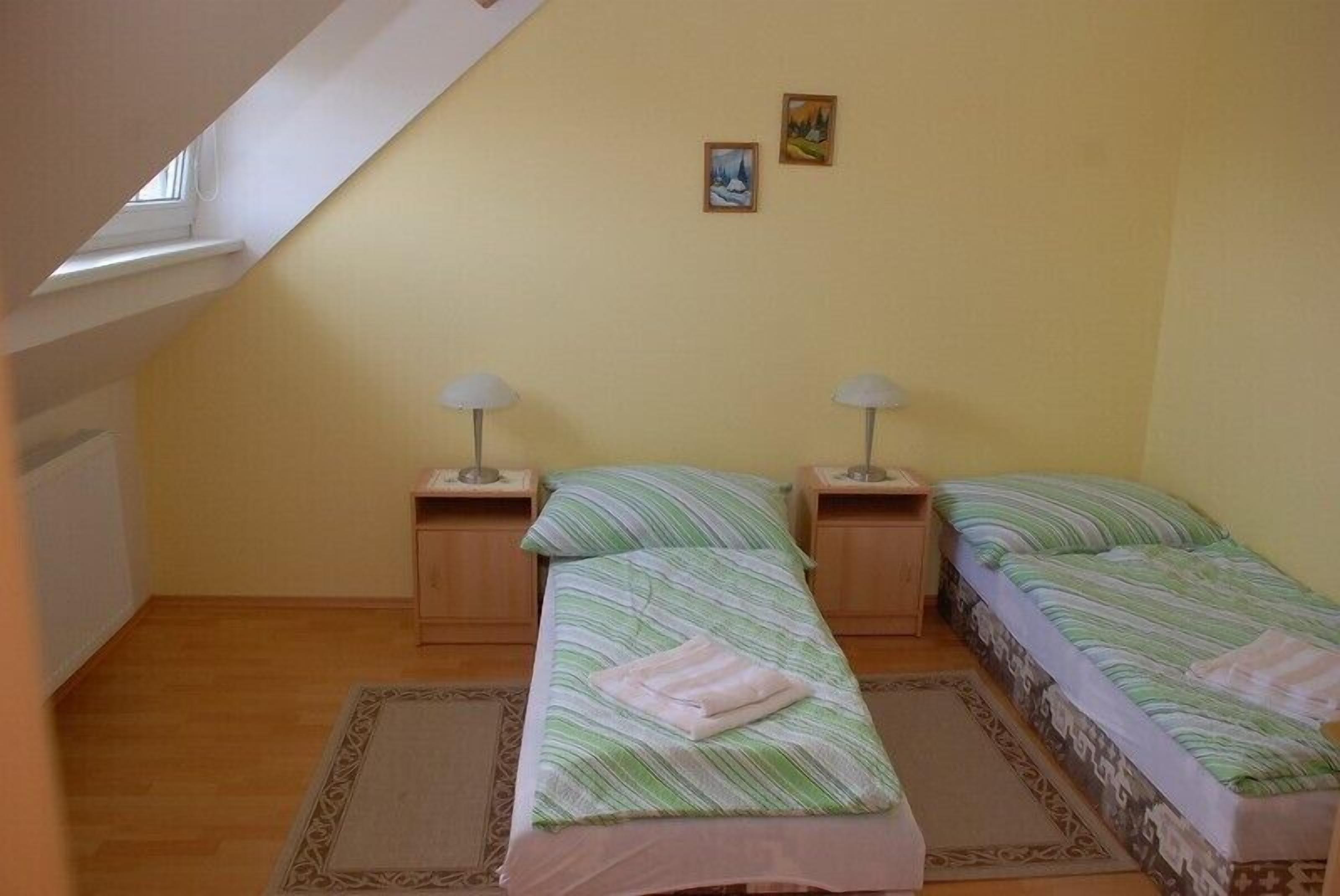 Ein kleiner, gemütlicher Raum mit zwei Einzelbetten, jeweils mit Nachttisch und Lampe. Das Zimmer hat eine schräge Decke mit einem Dachfenster, und die Wände sind hellgelb gestrichen.