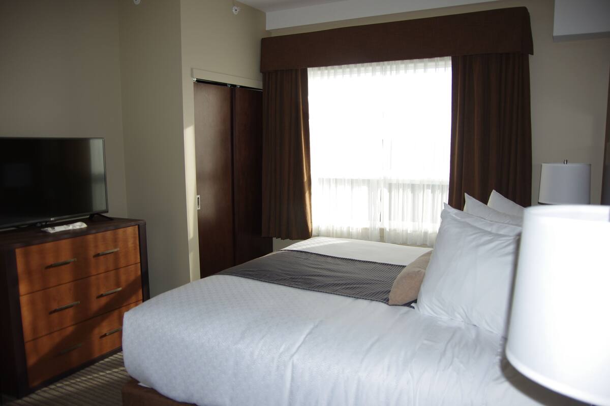 Ein Hotelzimmer mit Bett, Fernseher auf einer Kommode, Lampe und einem Fenster mit Vorhängen.