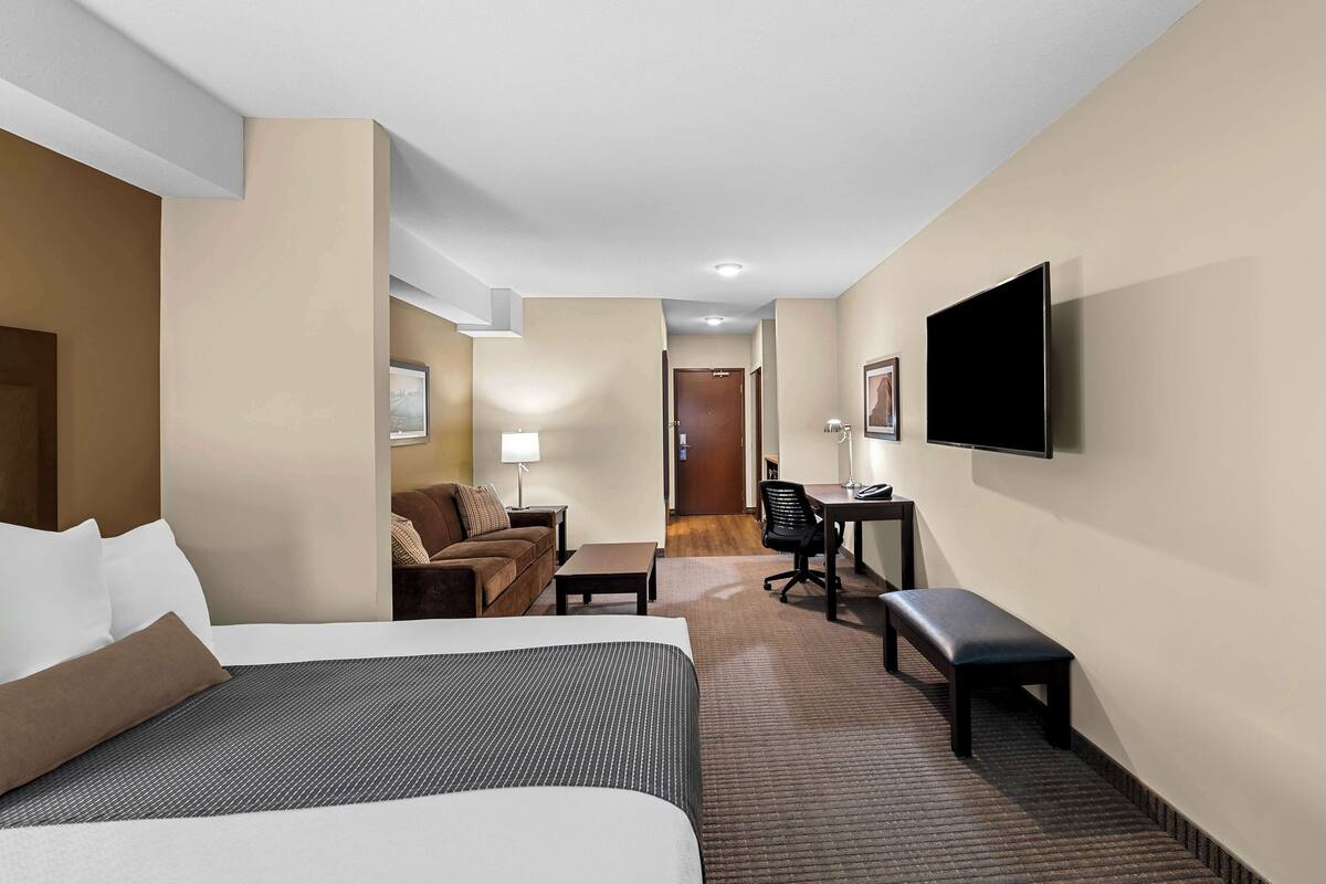 Ein Hotelzimmer mit Bett, Schreibtisch, Stuhl, Bank, Fernseher und einer Tür zu einem weiteren Raum.