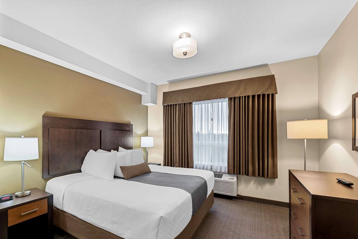 Ein Hotelzimmer mit einem großen Bett, zwei Nachttischen, einem Schreibtisch, einem Stuhl, einer Lampe, einem Spiegel und einem fenster mit Vorhängen.
