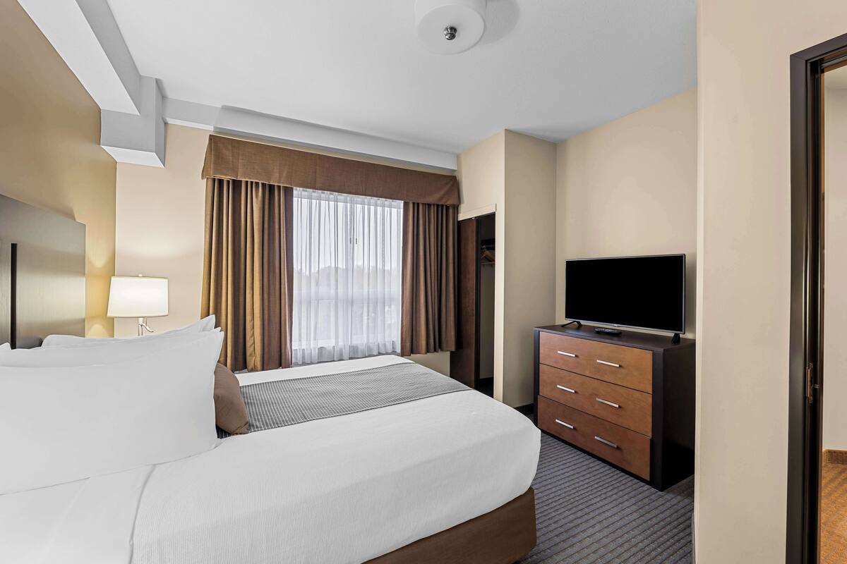 Ein Hotelzimmer mit einem großen Bett, einem Fernseher auf einer Kommode, einem Vorhangfenster und einer Lampe.