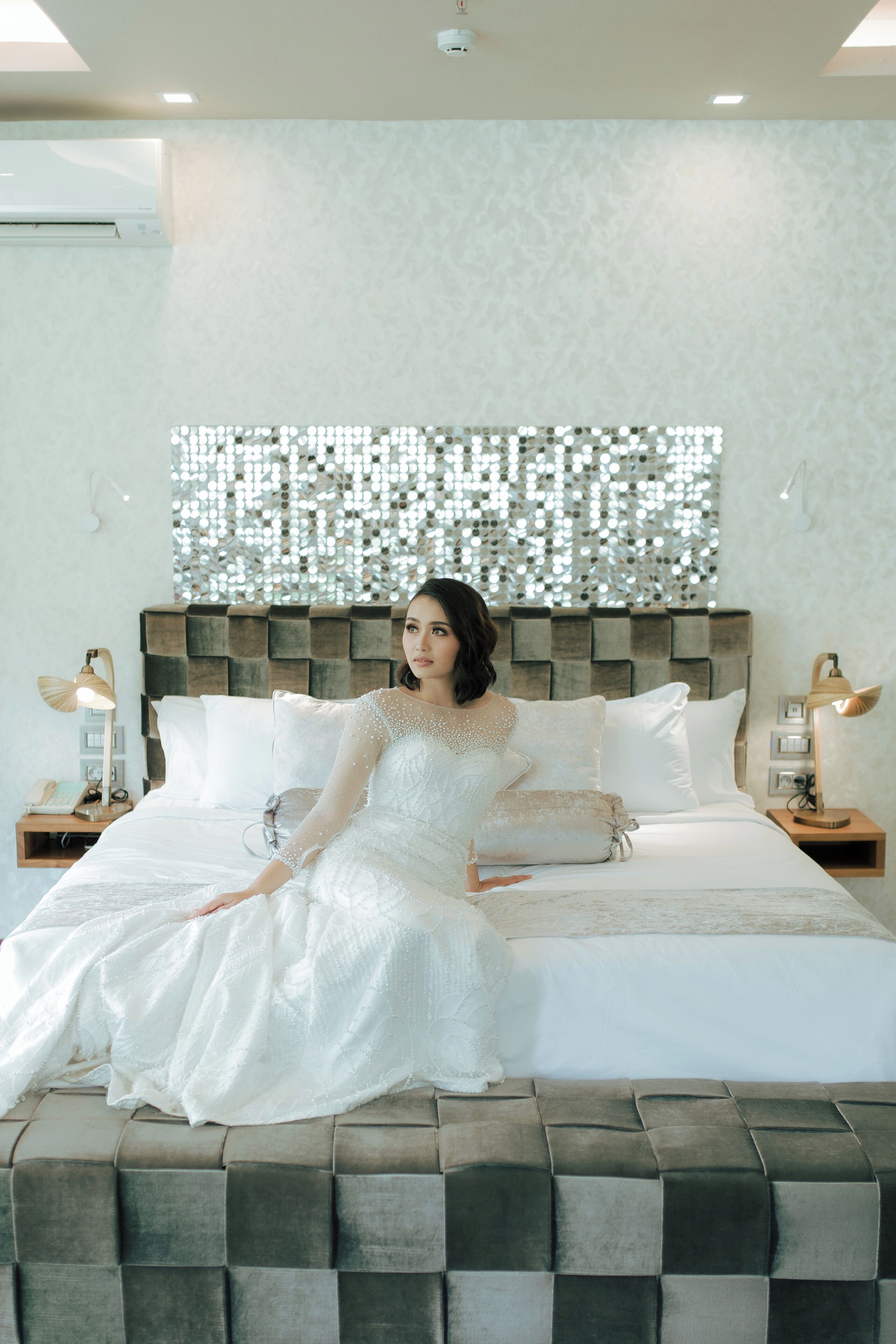 A bed with a decorative headboard, a bedside table with a lamp, and a wall with a reflective panel.