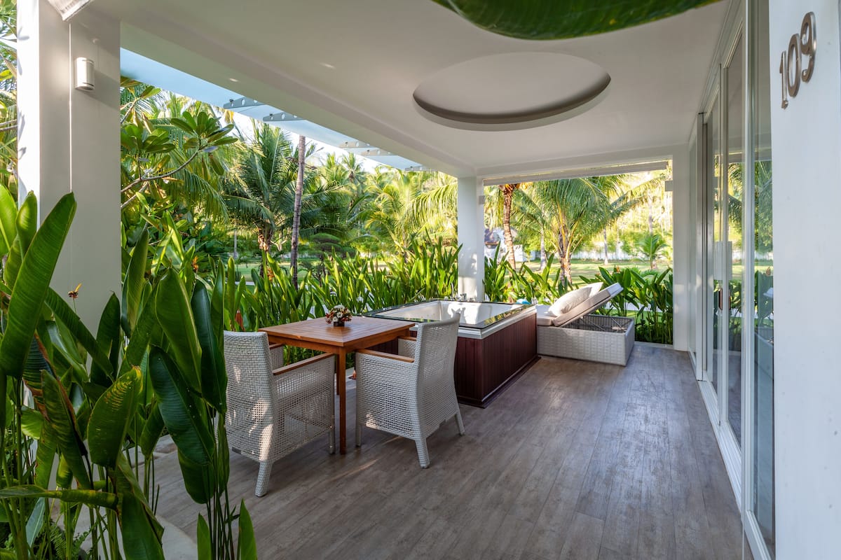 A modern outdoor lounge area with a wooden table, wicker chairs, a hot tub, and lush tropical plants.