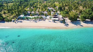 Eine Luftaufnahme eines Strandraises mit mehreren Bungalows über dem Wasser, einem Sandstrand und klarem türkisblauem Wasser.