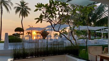 A modern outdoor pool area with a jacuzzi, surrounded by palm trees and umbrellas, during sunset.