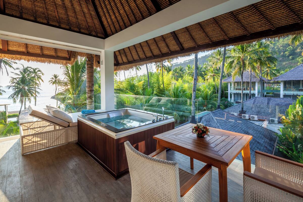 A modern room with a glass-enclosed hot tub, wicker furniture, and a view of lush greenery and a beach.