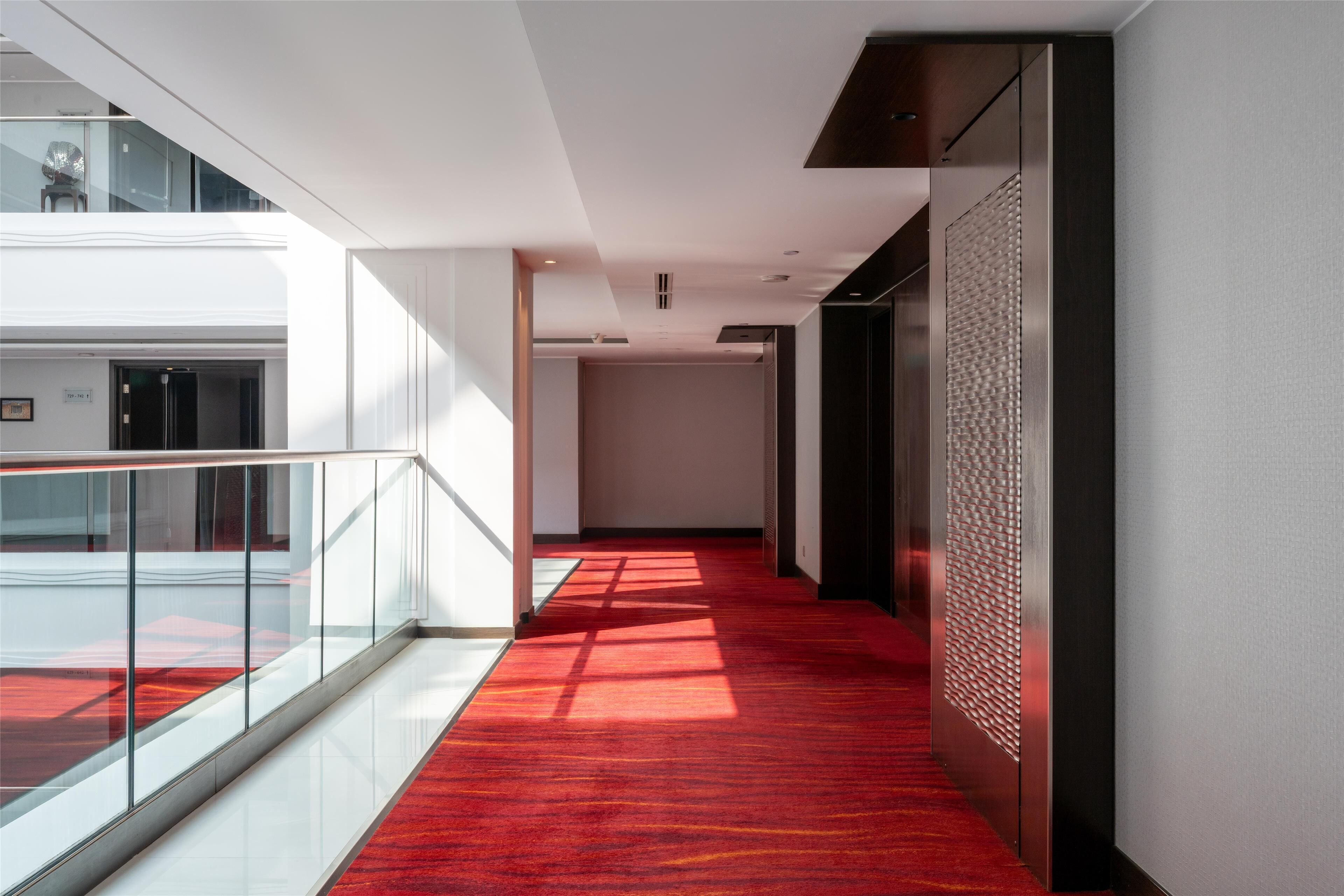 Un pasillo moderno de hotel con alfombra roja, barandillas de vidrio y pared texturizada.