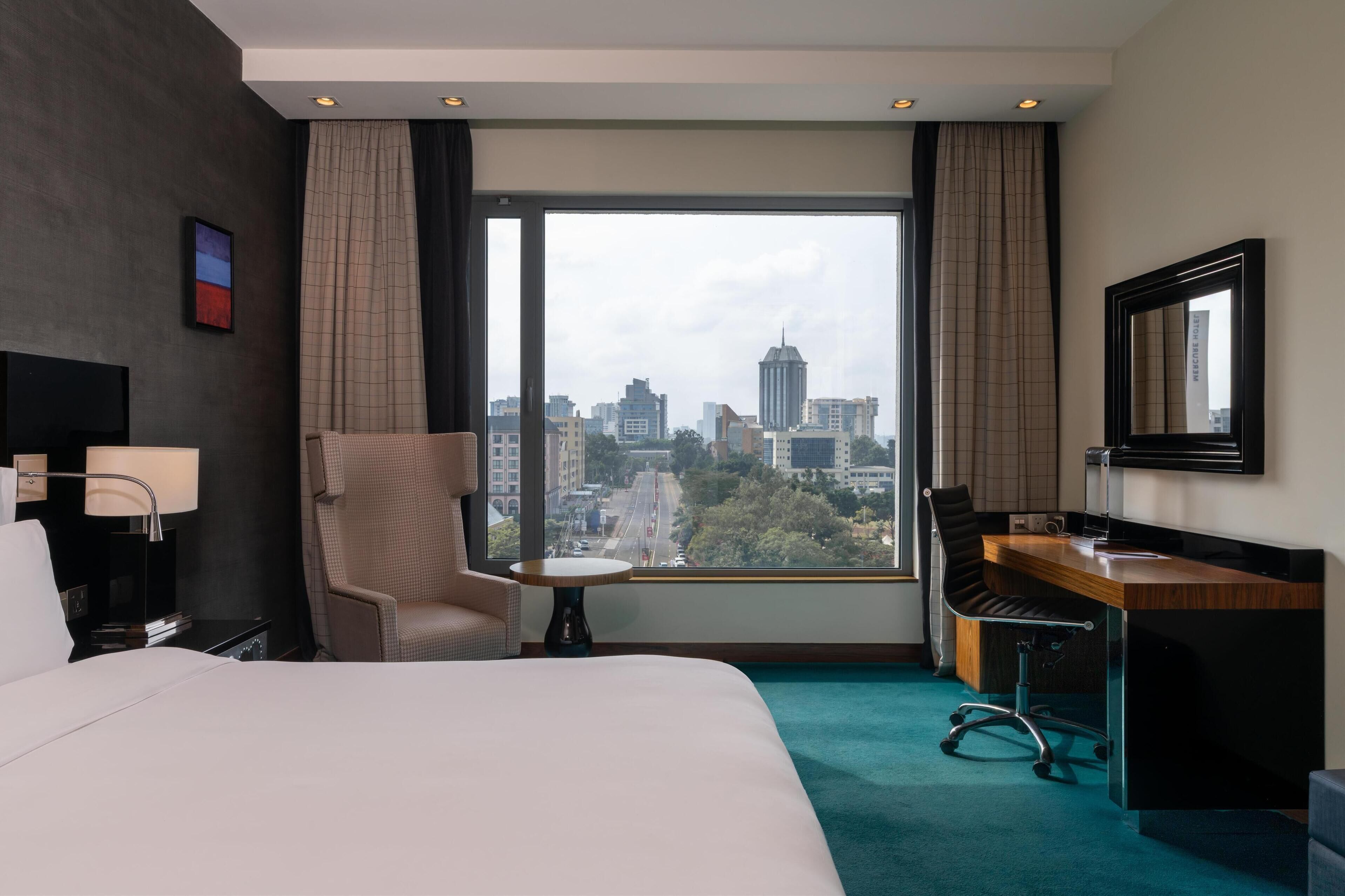 A hotel room with a large bed, a desk with a chair, a TV, and a view of a cityscape through a large window.