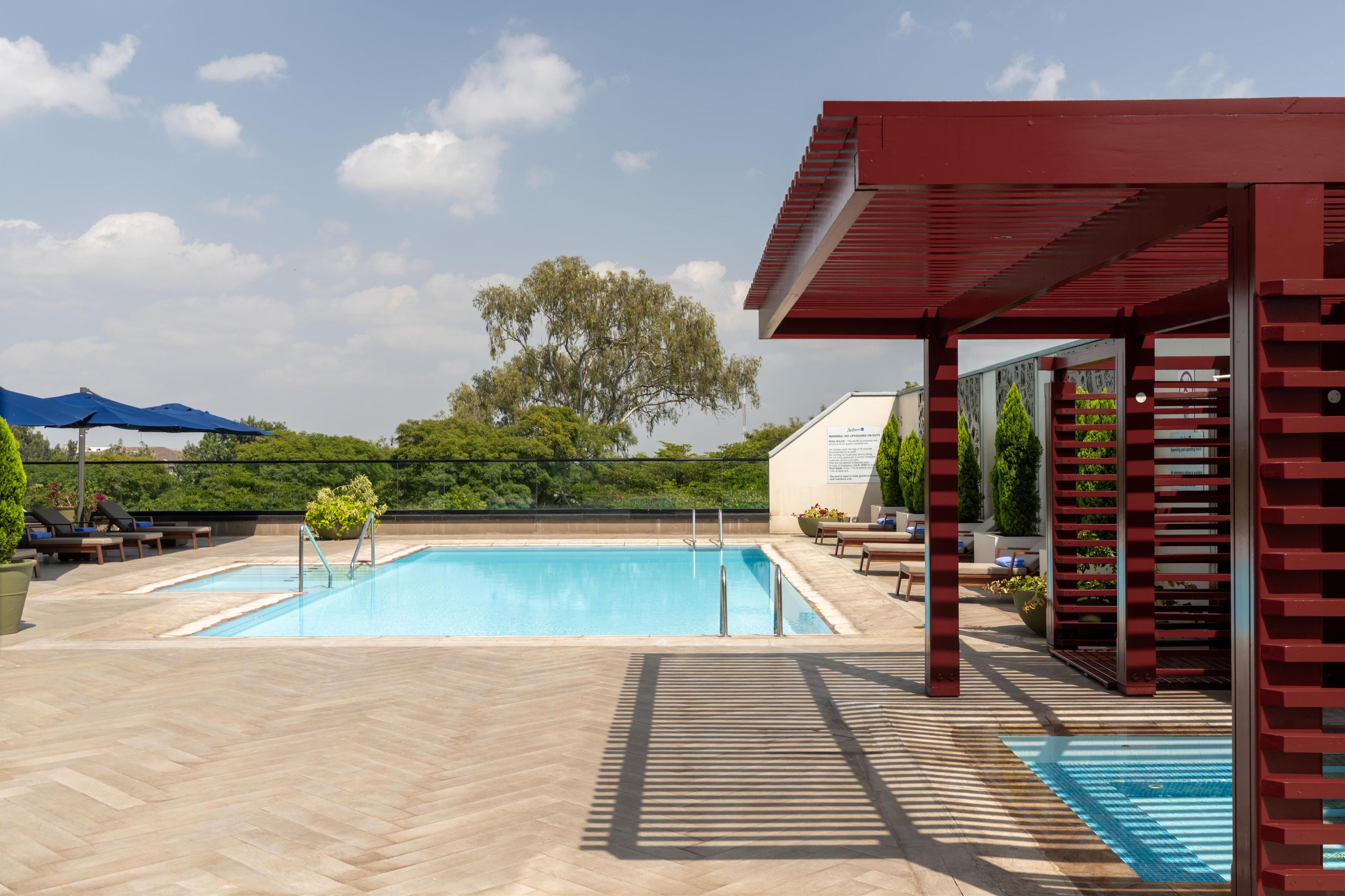 A swimming pool with a wooden deck, a covered seating area, and a view of trees and a building in the background.