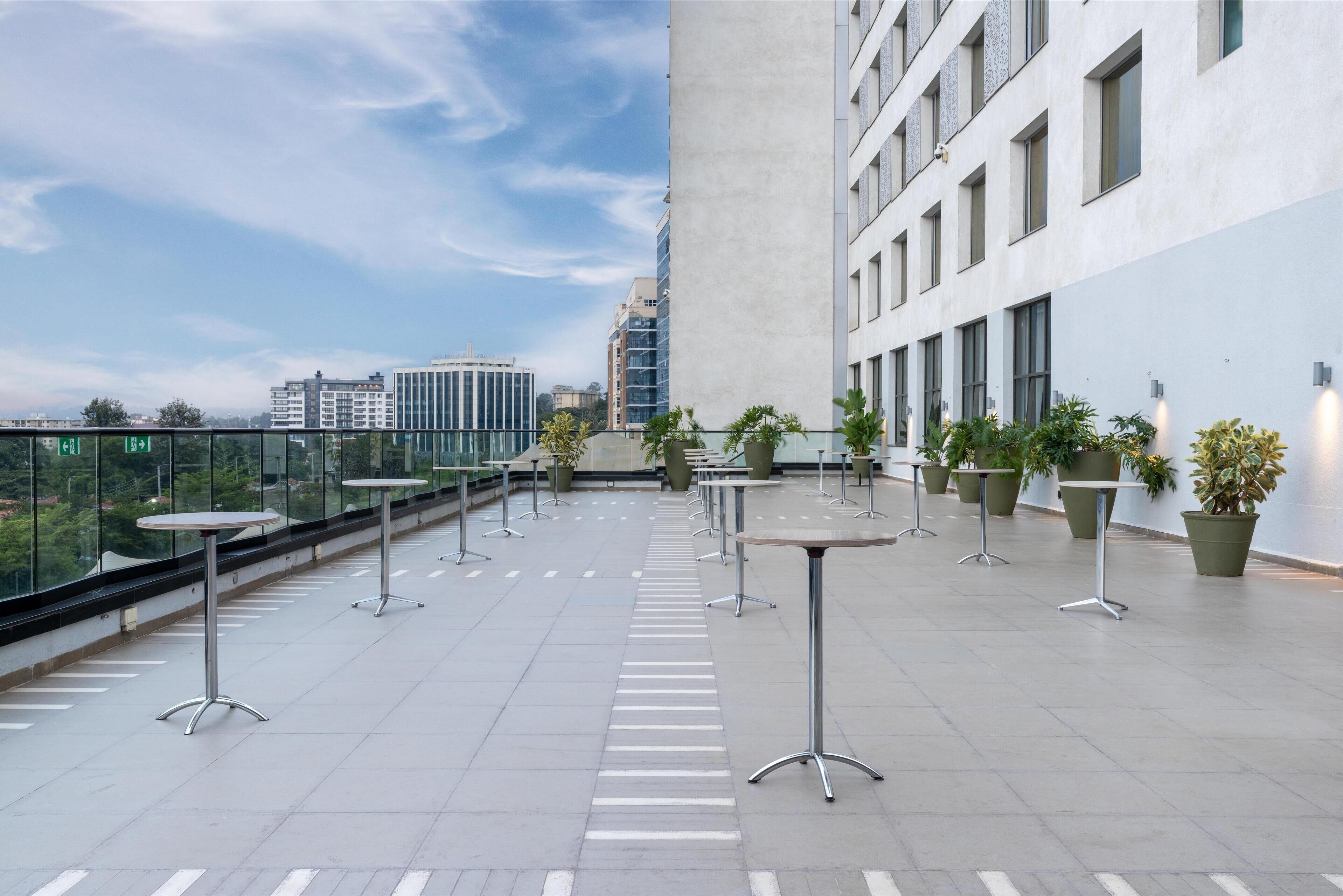 A rooftop terrace with tables and potted plants, overlooking a cityscape.