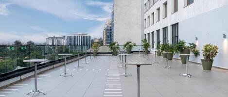 A rooftop terrace with tables and potted plants, overlooking a cityscape.