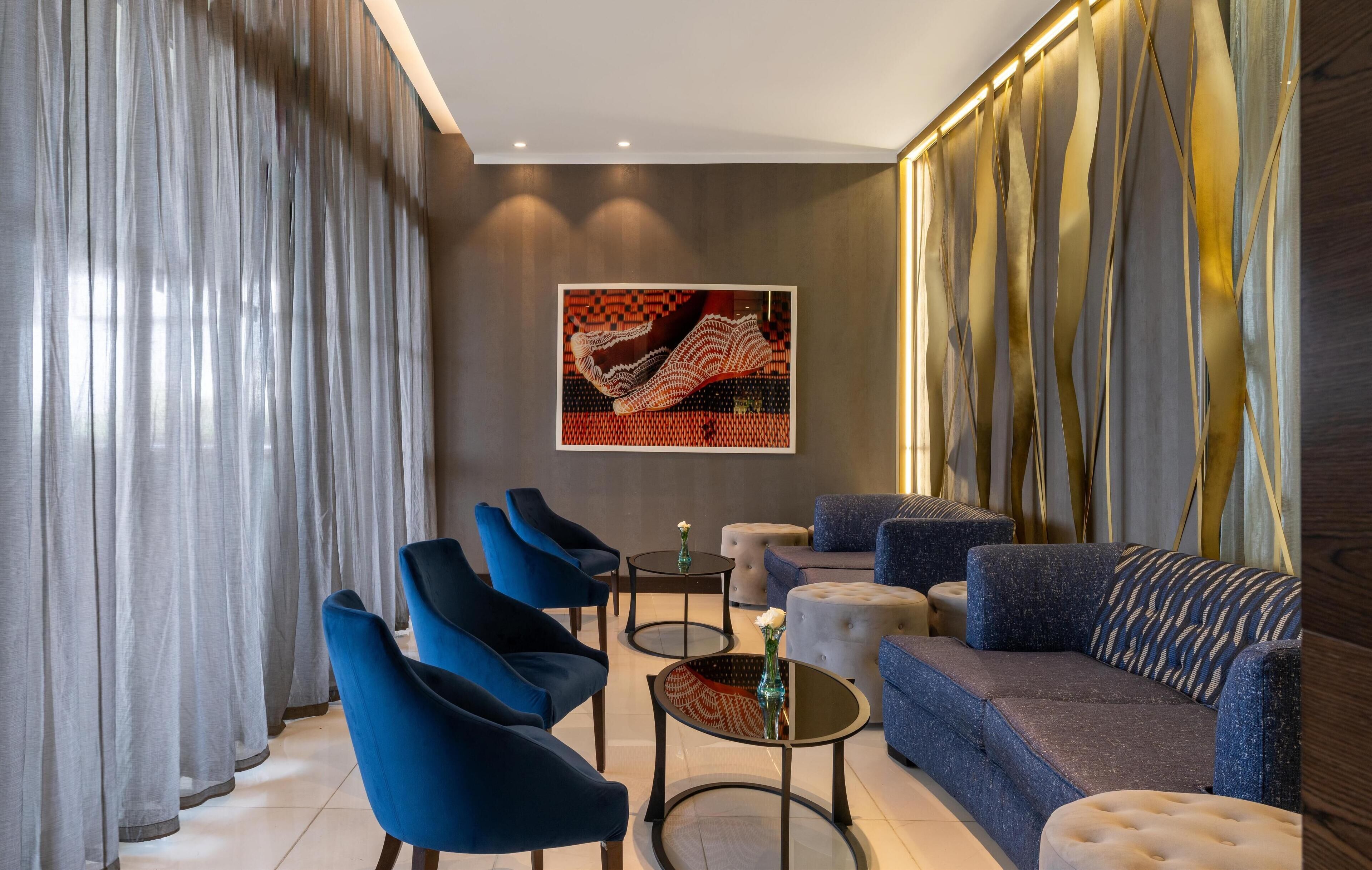 A modern living room with blue chairs, a sofa, and a coffee table. There is a large abstract painting on the wall and a window with curtains.