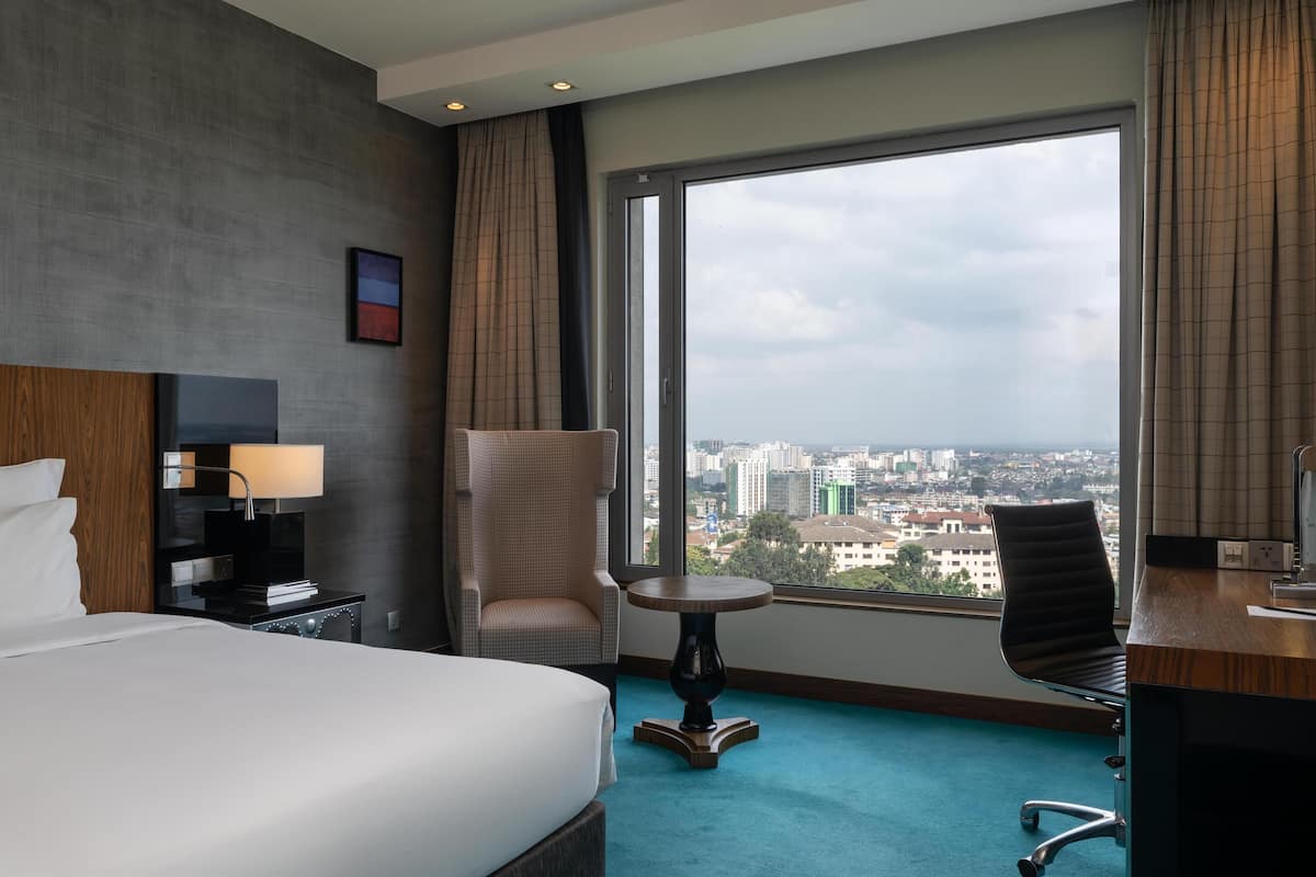 Quarto de hotel com uma cama grande, uma escrivaninha, uma cadeira, uma mesinha e vista para a cidade através de uma janela ampla.