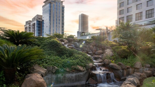 Een waterval in een aangelegde omgeving met grote rotsen en begroeiing, omringd door moderne gebouwen.