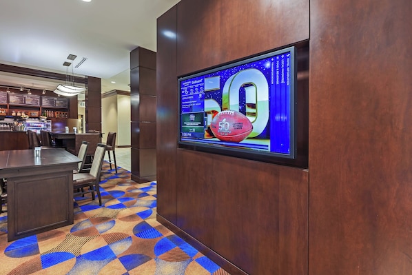 A hotel lobby with a large screen displaying a football game, a bar area, and a patterned carpet.